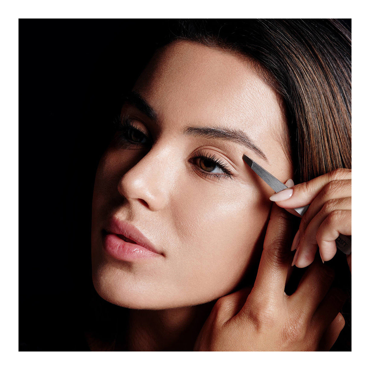 A person carefully uses RevitaLash Precision Tweezer to shape their arched brows against a dark background, focusing intently. The lighting highlights their smooth complexion and well-groomed features.