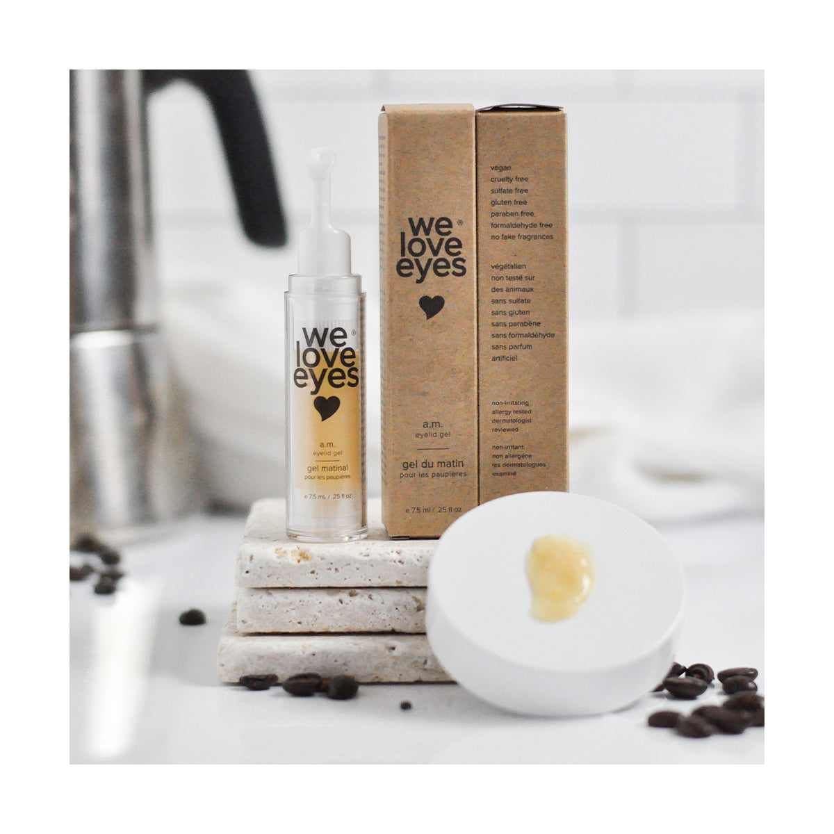 A 7.5ml bottle of We Love Eyes - AM Eyelid Gel sits on stacked stone tiles, promoting hydrating eye care. Behind it, a brown cardboard box with the same branding is surrounded by scattered coffee beans and a blurred faucet.