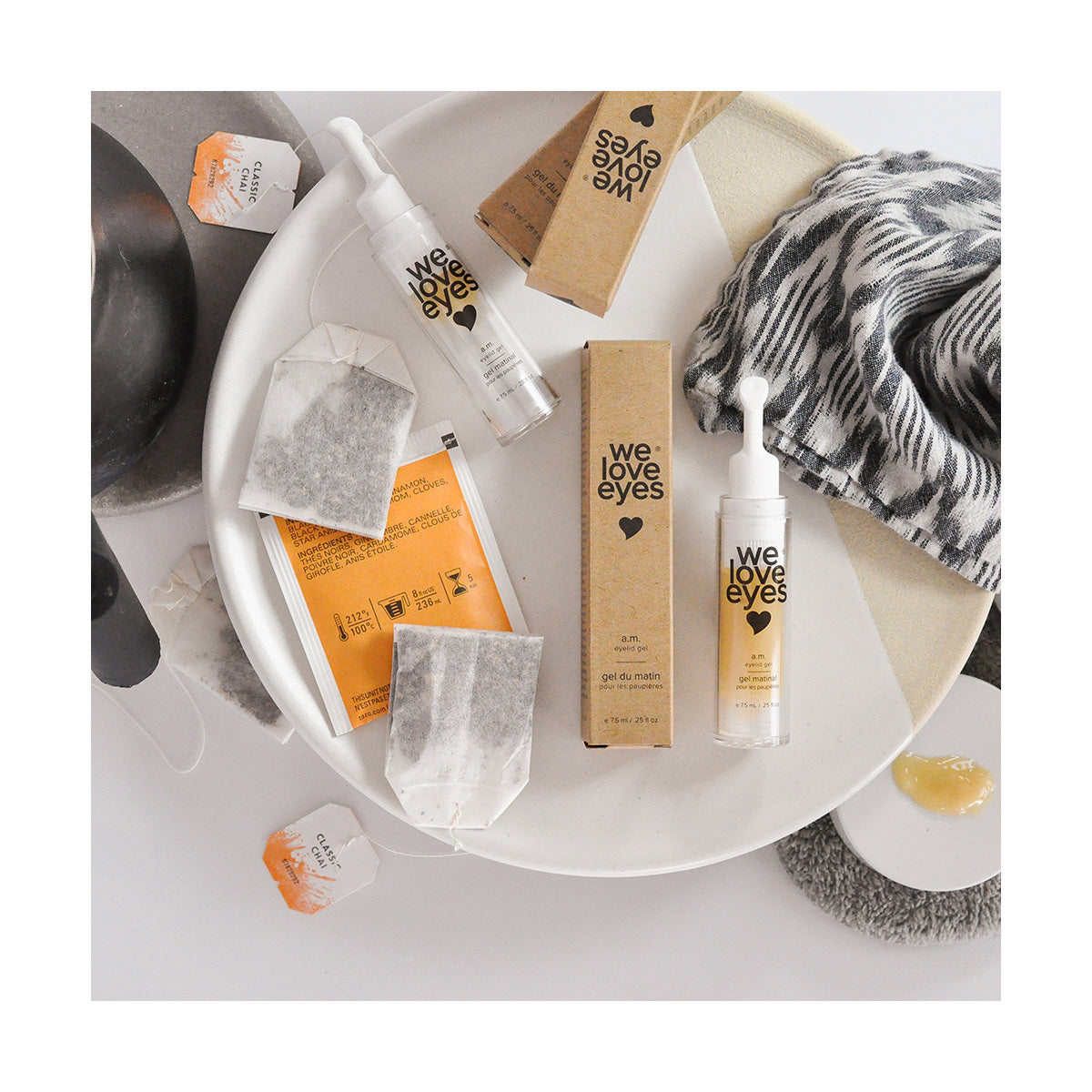Flat lay featuring We Love Eyes - AM Eyelid Gel with its packaging on a white plate, alongside tea bags, a striped towel, and a small cloth with a yellow substance; this arrangement showcases hydrating eye care for optimal meibomian gland health.
