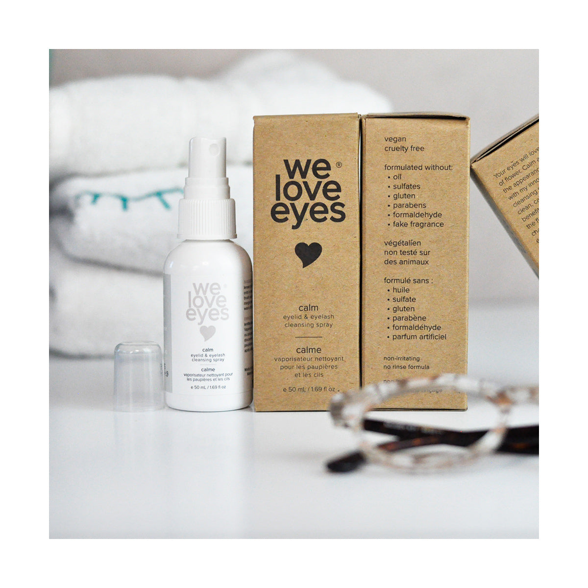 A We Love Eyes Calm Eyelid & Eyelash Cleansing Spray with HypoChlorous stands next to a brown box, showcasing its chamomile and hypochlorous acid blend. In the background, eyeglasses rest on white towels.