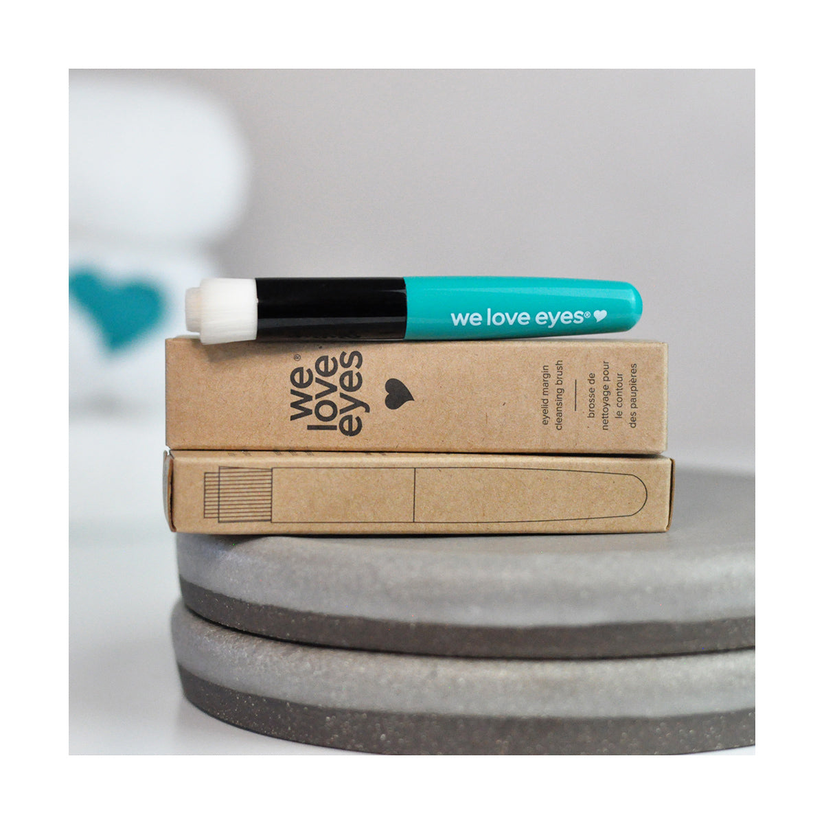 A teal We Love Eyes Eyelid Margin Cleansing Brush, ideal for eyelid cleansing, is placed on two brown boxes with We Love Eyes and a heart printed on them, resting on a gray circular base. A blurred blue shape in the background provides an artistic accent.