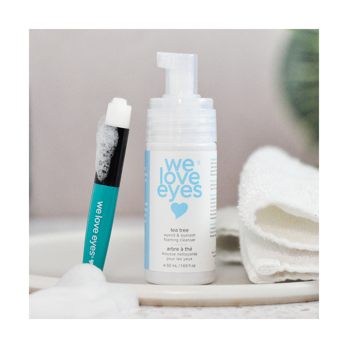 A bottle of We Love Eyes tea tree eyelid and eyelash foaming cleanser, designed for optimal lash follicle health, is paired with the We Love Eyes - Eyelid Margin Cleansing Brush, featuring foam on its tip. A soft towel appears blurred in the background.