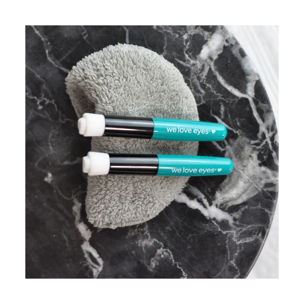 Two teal We Love Eyes tubes with white tips, designed to support lash follicle health, rest on a gray textured pad and sit elegantly on a dark marble surface.