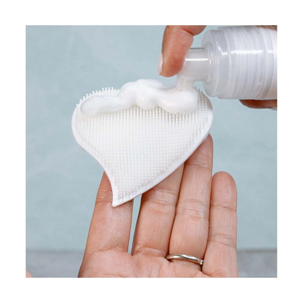 A person dispenses foam from the We Love Eyes cleanser onto a white, heart-shaped We Love Eyes Eyelid Scrub Mitt held in their hand, set against a light green background. A silver ring embellishes their ring finger, adding elegance to the scene.