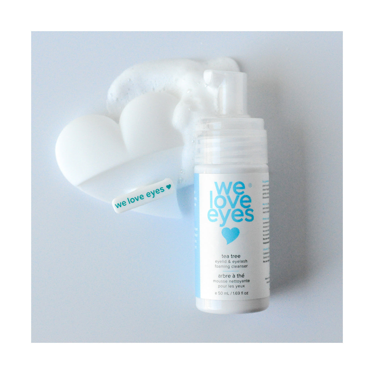 A We Love Eyes tea tree eyelid foaming cleanser sits beside heart-shaped foam, paired with a We Love Eyes Eyelid Scrub Mitt. The label is white with blue text, and a small sticker reads we love eyes alongside a green heart.