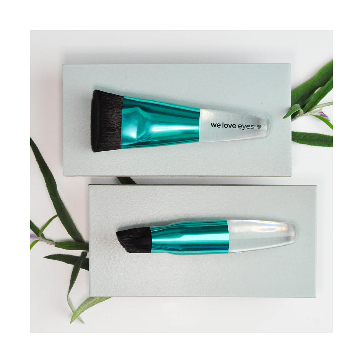 Two makeup brushes with teal metallic handles rest on gray rectangular platforms. The top brush, the We Love Eyes Lashfull Thinking brush, is vegan and cruelty-free and promotes eyelid margin health. Green leaves are elegantly arranged around the platforms for decoration.