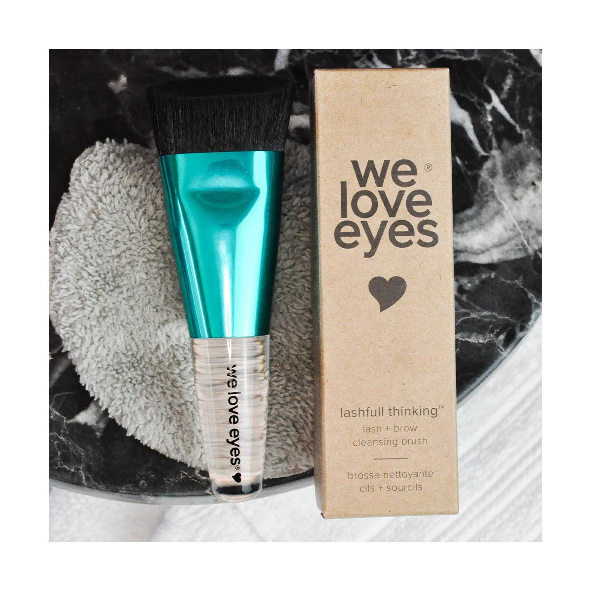 The We Love Eyes - Lashfull Thinking lash and brow cleansing brush, with a green handle, rests on a black-and-white marble surface to promote eyelid margin health. The vegan, cruelty-free packaging is visible beside a textured gray cloth partially peeking out.