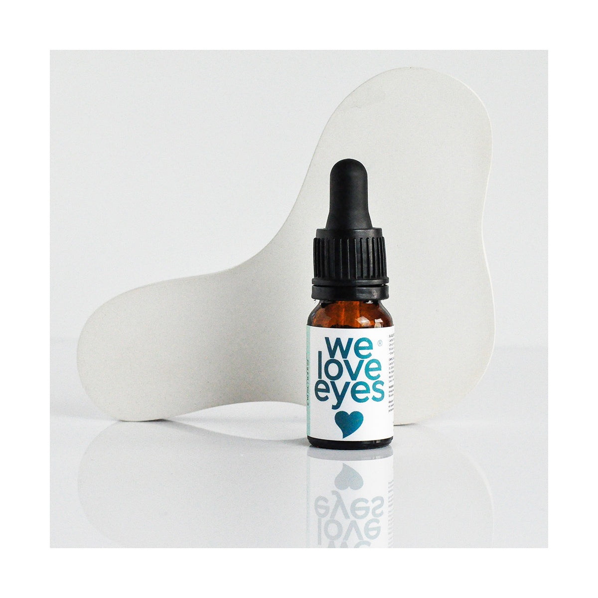 A 10mL bottle of We Love Eyes Lashfull Thinkin Lash + Brow Follicle Oil rests before a curved white abstract shape, its reflection gleaming on the glossy surface below. The amber bottle features a dropper and is infused with ophthalmic-grade castor oil.