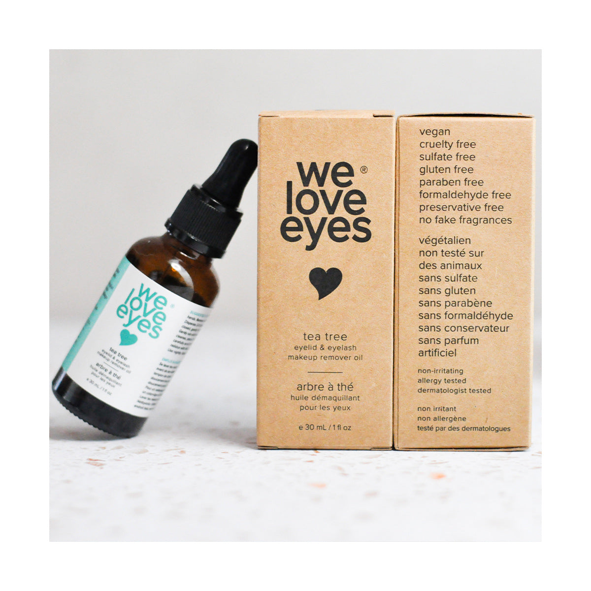 A 30mL glass dropper bottle labeled We Love Eyes is next to a brown box promoting its makeup remover oil with tea tree oil. Its vegan, cruelty-free, and contains no sulfates, gluten, parabens, preservatives, or artificial fragrances.