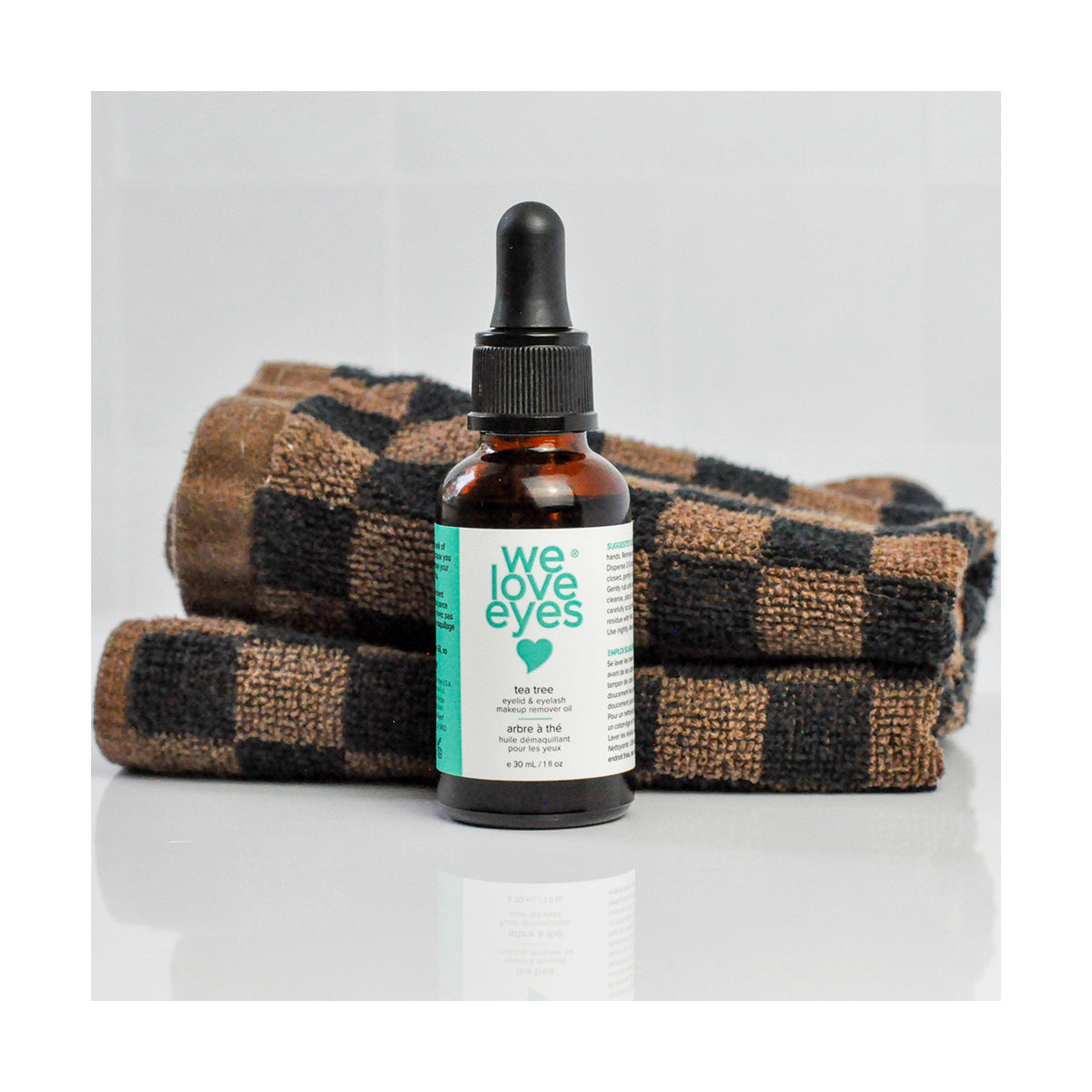 A We Love Eyes bottle with a turquoise label and dropper cap, containing 30mL of all-natural tea tree makeup remover oil, is set against a white background before brown and black checkered towels.