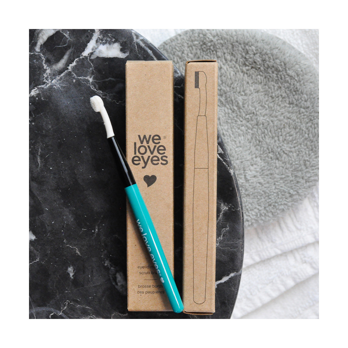Two brown boxes labeled We Love Eyes accompany a We Love Eyes Eyelid Margin Scrub Brush with a turquoise handle on dark marble. A gray towel and white cloth in the background highlight a focus on meibomian gland health and eye care.