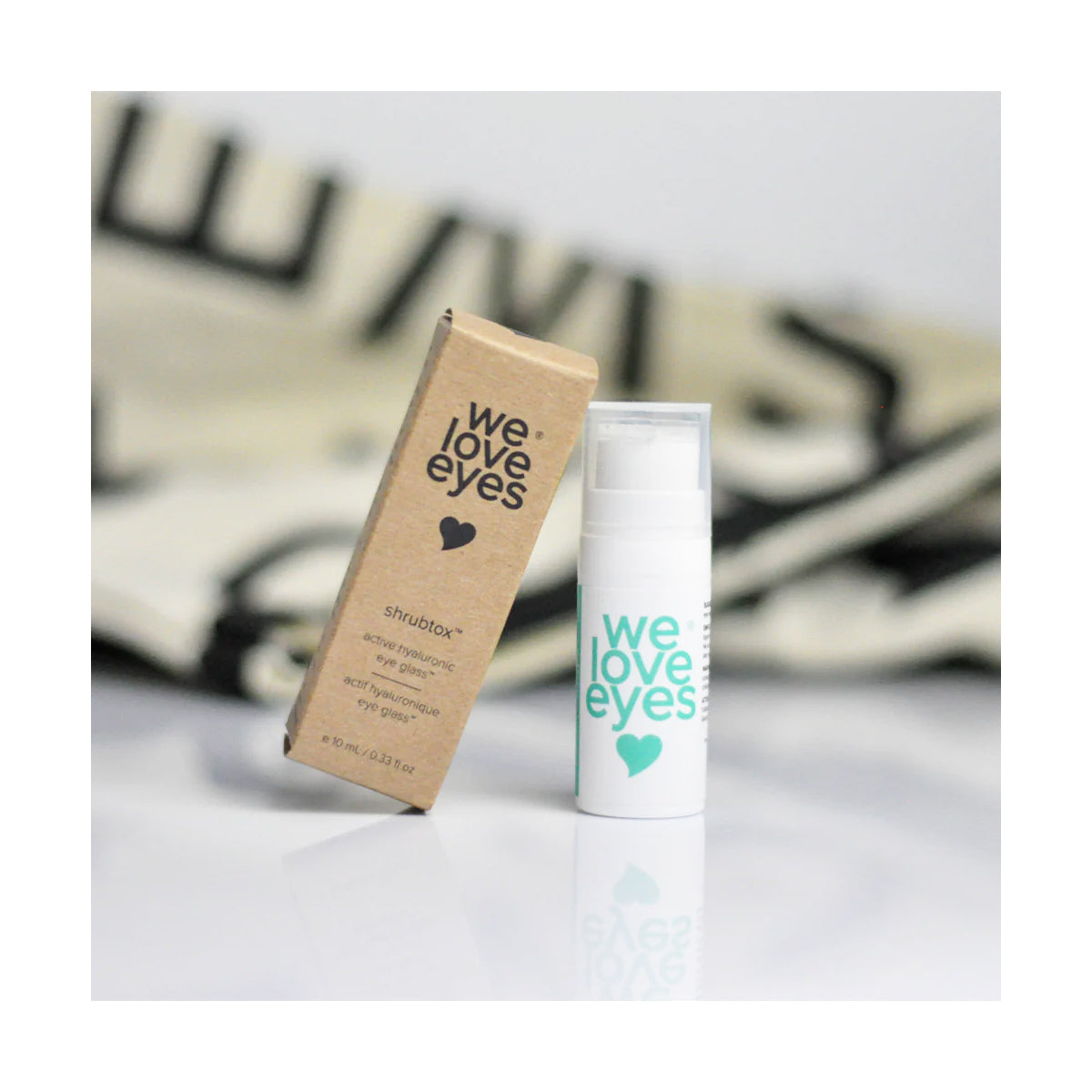 A brown box labeled We Love Eyes is next to a white bottle featuring a teal heart and the same text, advertising We Love Eyes Shrubtox Active Hyaluronic Eye Glass (10mL Bottle) for eyelid skin barrier care. The background has blurred black and white patterns.
