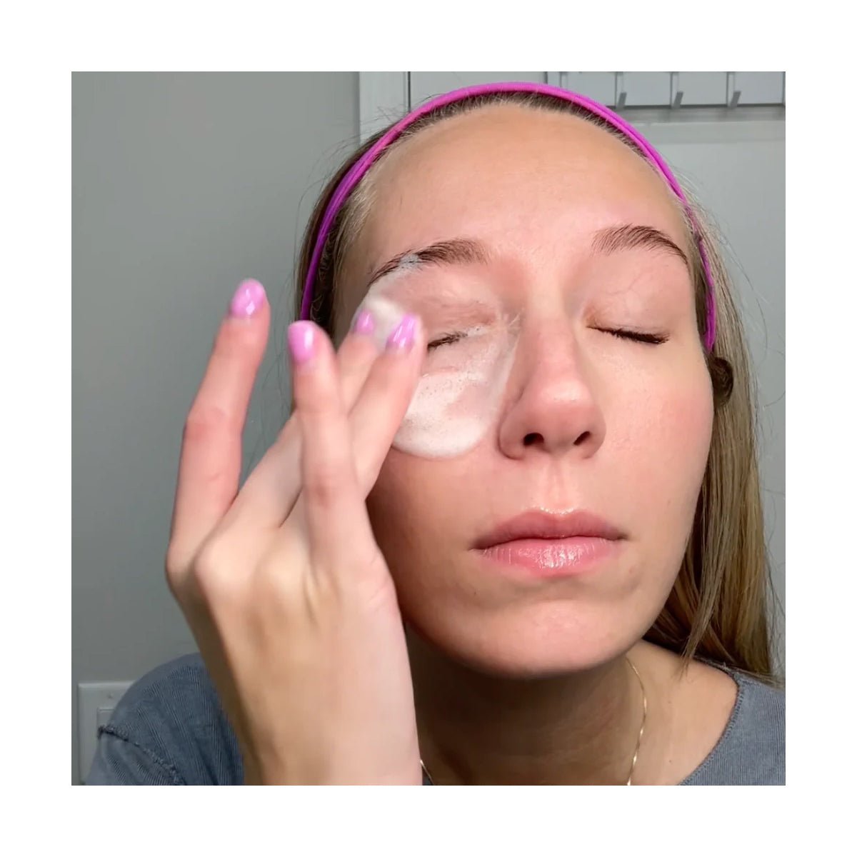 A person with closed eyes gently applies We Love Eyes SuperGLA eyelid & eyelash foaming cleanser (50mL) using their fingers. Wearing a pink headband and light pink nail polish, they stand against a neutral wall, ensuring gentle care for dry eyes without irritation.