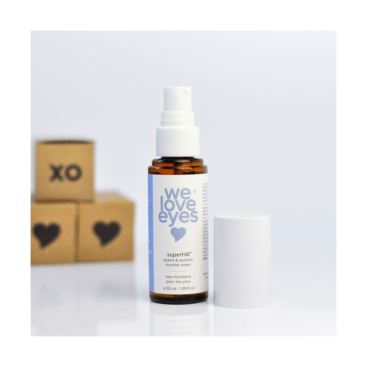 A brown bottle of We Love Eyes superHA Eyelid & Eyelash Micellar Water Make-up Remover (50mL) with its white cap removed stands center stage. Behind it, blurred blocks featuring a heart and XO create a bright, minimalistic scene.