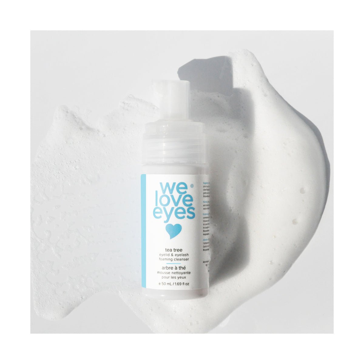 The We Love Eyes Tea Tree Eyelid & Eyelash Foaming Cleanser 25mL Mini Size from We Love Eyes is showcased on a white, foamy background. The clean white bottle with a blue label and pump dispenser, infused with tea tree oil, creates a gentle contrast that highlights its soothing effect.