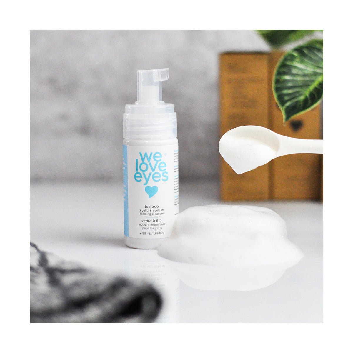 A reflective surface displays a white pump bottle with blue text, We Love Eyes: Tea Tree Eyelid Foaming Cleanser, alongside a spoon of foam and a green leaf. The brown box in the background emphasizes its plant-based formula enriched with tea tree oil.