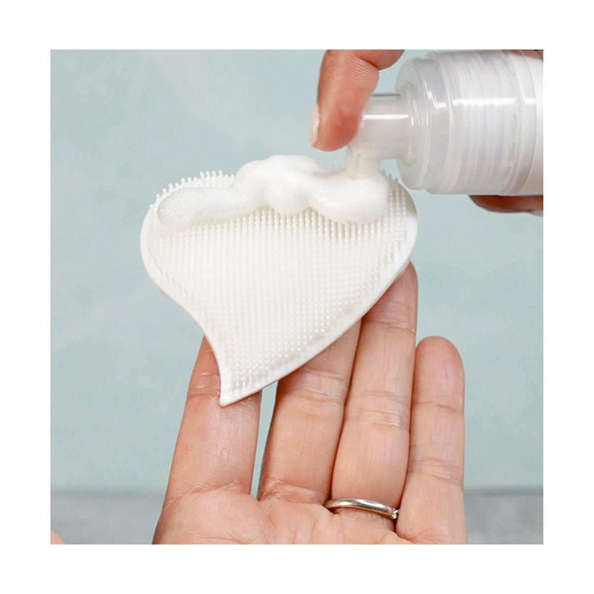 A hand holds a white, heart-shaped cleansing pad with textured bristles, as We Love Eyes: Tea Tree Eyelid Foaming Cleanser is dispensed onto it. The background is a soft blue.