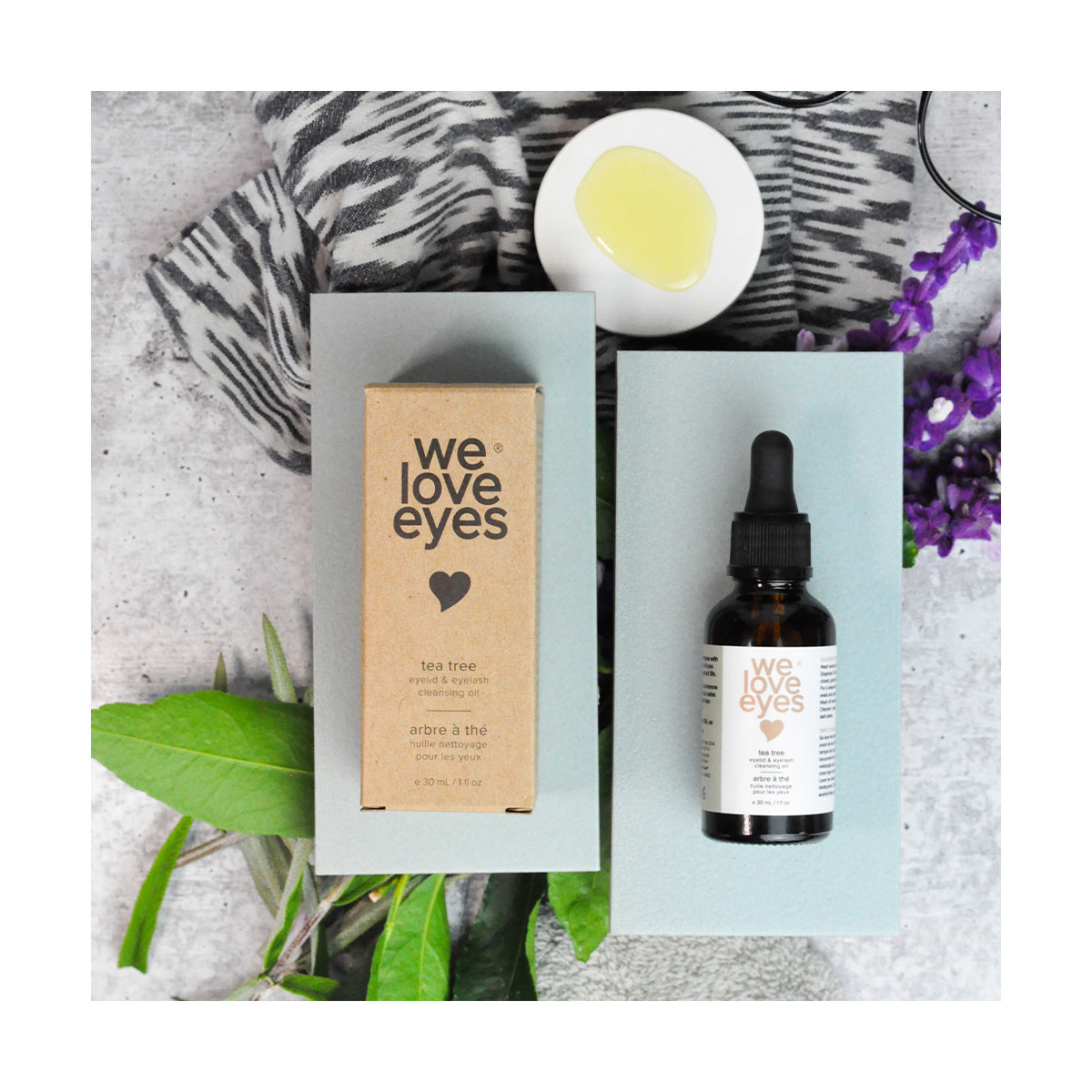 A box labeled We Love Eyes and a bottle of We Love Eyes- All Natural Tea Tree Eyelid Cleansing Oil (30ml) rest elegantly on gray surfaces. Nearby, a small dish with oil, a patterned cloth, green leaves, and purple flowers create a serene setting that nurtures eyelid skin and Meibomian oil glands.