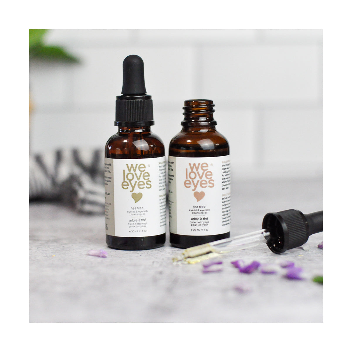 Two amber We Love Eyes dropper bottles, one open and one sealed, sit on a gray surface with purple flowers. These 30ml All Natural Tea Tree Eyelid Cleansing Oils showcase the benefits of Tea Tree Oil for eyelid skin.