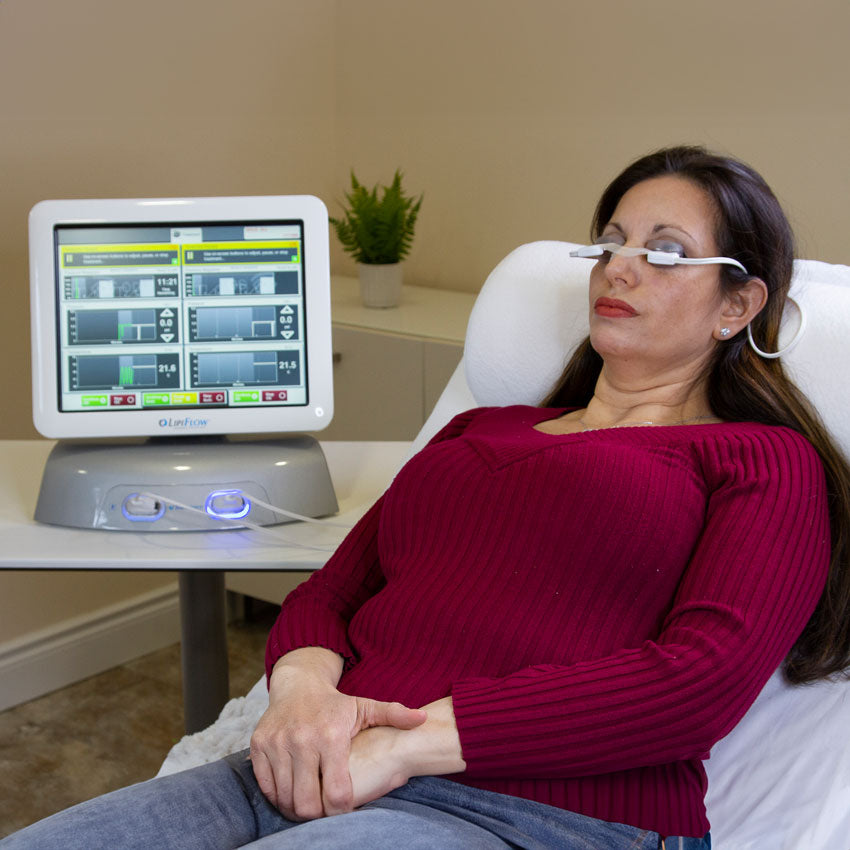 A woman in a red sweater reclines in a chair with electrodes on her forehead, connected to a device next to her. The device has a screen displaying graphs and data. A small plant is in the background.