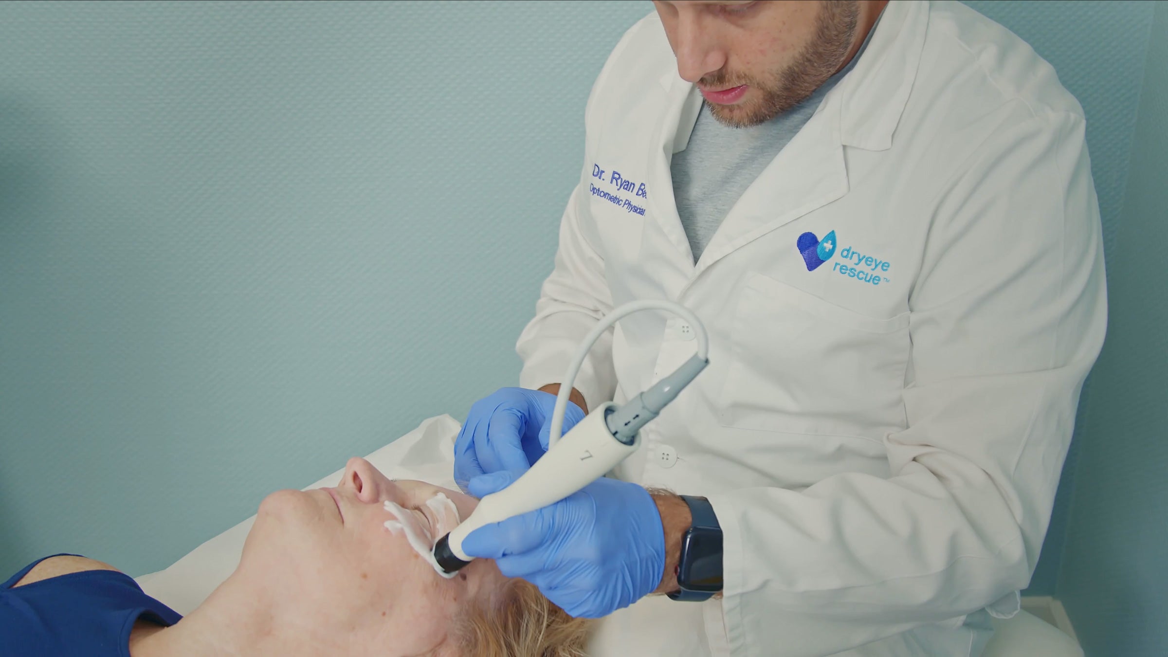 A healthcare professional in a white coat and blue gloves uses a medical device on a patients face. The patient is lying down and has a relaxed expression. The setting appears to be a clinical or treatment room.