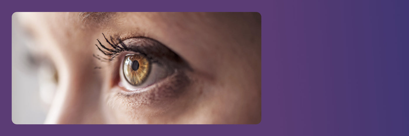 Close-up image of a persons eye gazing to the side, with long eyelashes and a detailed reflection in the iris. The background is a gradient of purple shades.