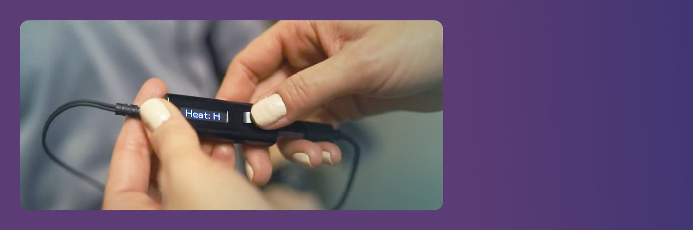 Hands holding a small black electronic device with a display showing the text Heat: H on a blue background. The device appears to have wires attached. The background is a gradient of purple shades.