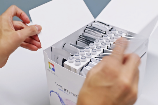A person is opening a white box labeled Inflamma containing multiple test tubes with grey caps. The box features a colorful logo with the name Quidel on the side. The persons hands are visible, handling the box.