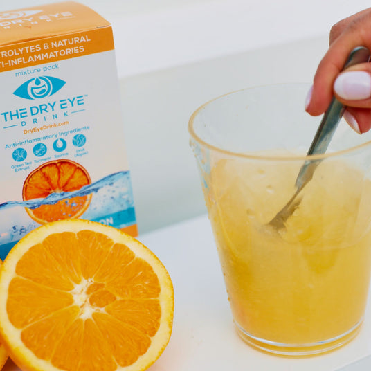 A hand stirring a yellow drink in a clear plastic cup with a spoon. Next to it, a sliced orange and a box labeled The Dry Eye Drink featuring electrolytes and natural anti-inflammatories. The packaging shows an orange graphic.