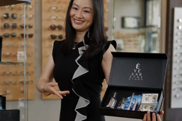 A woman in a black dress with a wavy white trim stands in an eyewear store, smiling. She is holding and pointing to a black box containing beauty products, including bottles and a mask. Eyewear displays are visible in the background.
