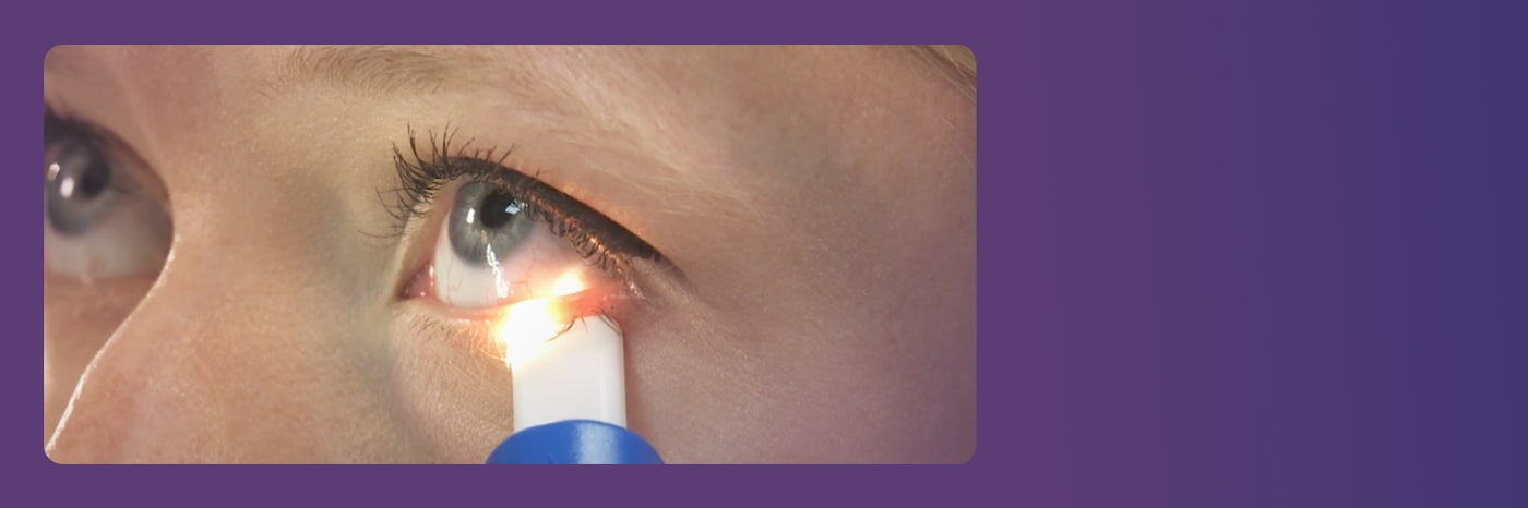 Close-up of a person using an eye device with a small light aimed at their left eye. The background is a gradient of purple shades.