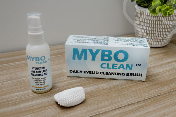 A bottle of Mybo Clean eyelid and lash cleansing gel, a cleansing brush, and the products packaging sit on a wooden surface. A small potted plant is in the background.
