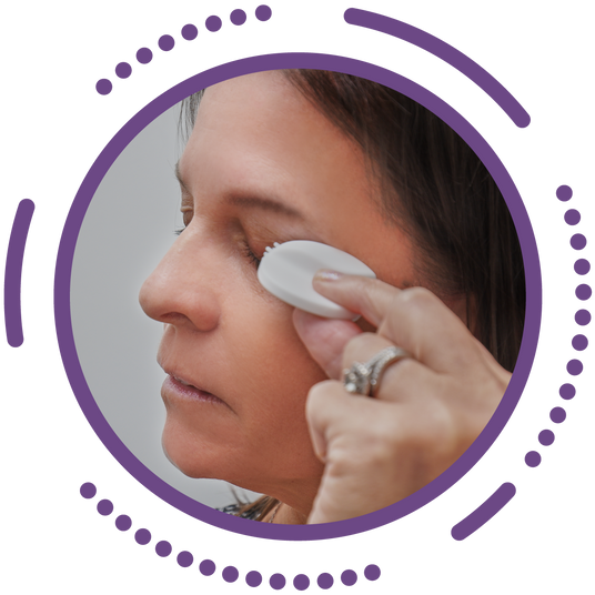 A woman gently applies an eyelid wipe to her closed eye. Her other eye remains closed, and she wears a silver ring. The image is framed by a circular, purple dotted border.