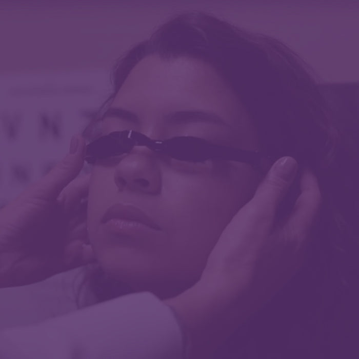 A person wearing dark, wraparound glasses is having them adjusted by someone elses hands. The image has a purple filter. The background is blurred and indistinct.
