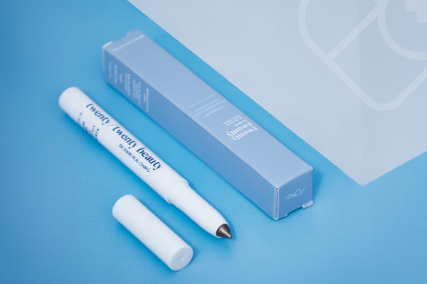 A white eye cream applicator with a silver tip and its cap removed lies next to its light blue box packaging on a blue surface. The text on the box and applicator is visible but not fully clear.