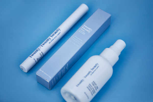 A set of beauty products on a blue background, including a white spray bottle, a slim cylindrical container, and a rectangular box, all labeled twenty/twenty beauty in blue text.