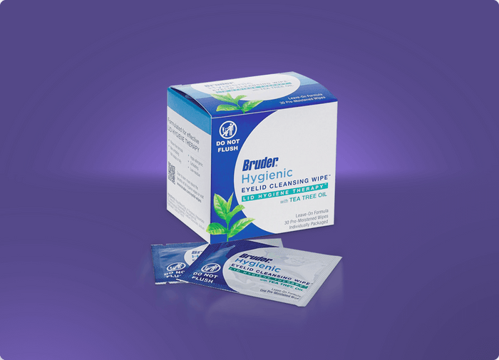 A box of Bruder Hygienic Eyelid Cleansing Wipes against a purple background. The box is white and blue with tea tree oil indicated. Two individual wipe packets are displayed in front of the box.