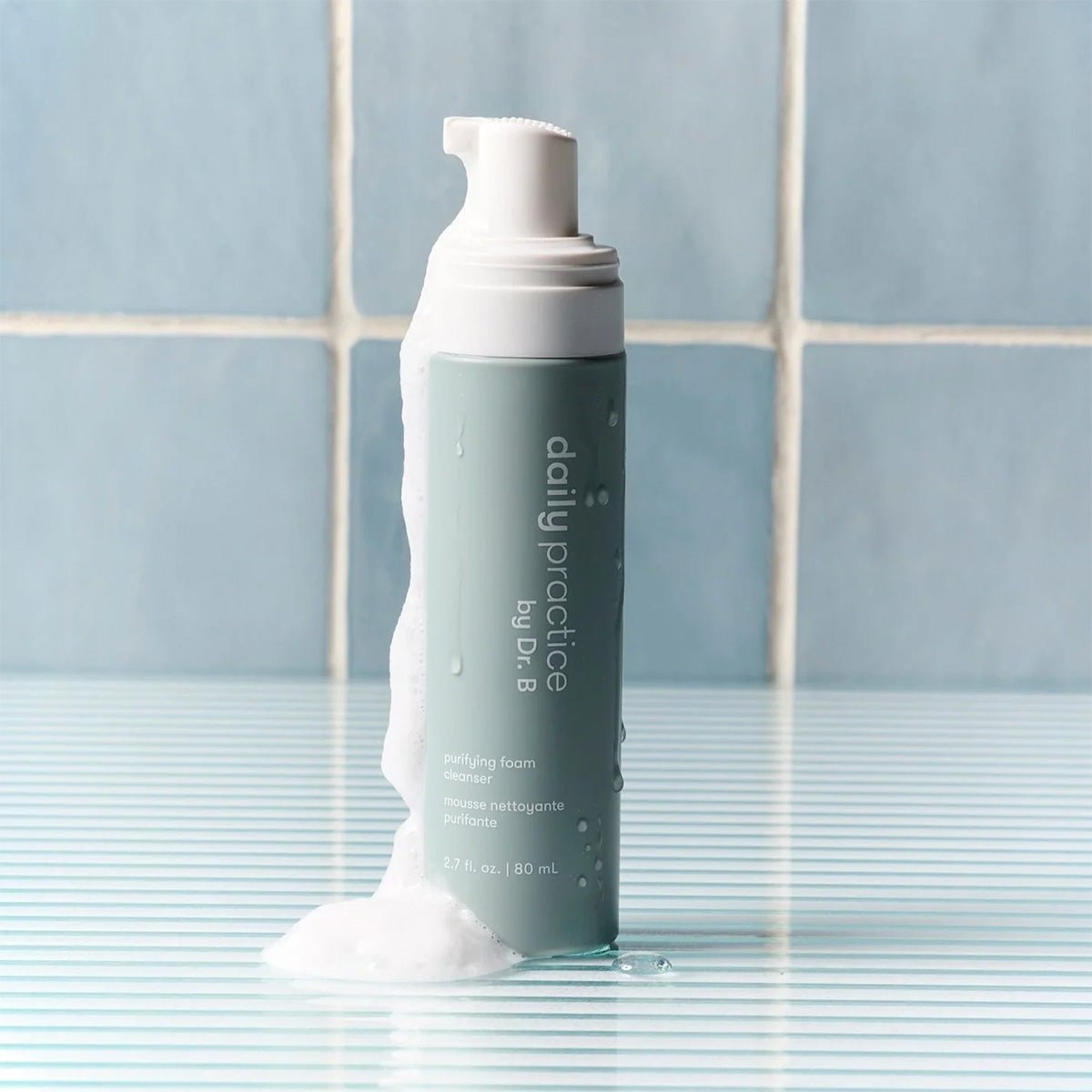 A light green bottle of Daily Practice Purifying Foam Cleanser, designed for eyes and lashes, sits on a striped surface with a tiled background. Foam overflows from the top, highlighting its gentle and hydrating formula for sensitive eyes.