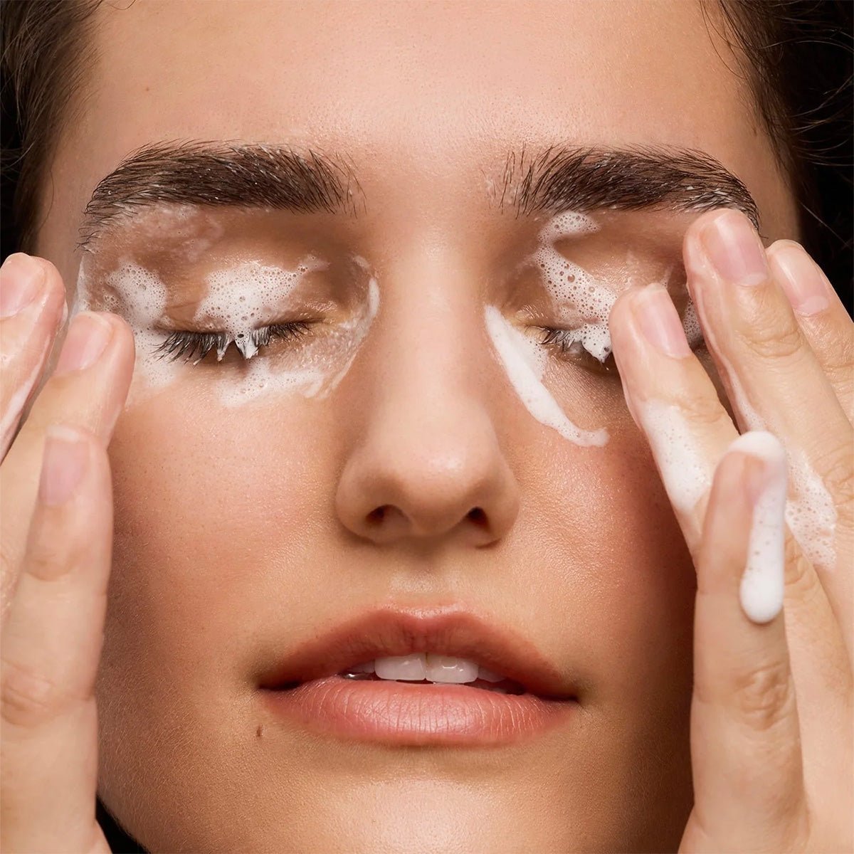 A person gently rubs their closed eyes with Daily Practice Purifying Foam Cleanser, forming a creamy lather. Skin appears smooth and well-hydrated, ideal for sensitive eyes and lashes. Lips are slightly parted, with emphasis on the eye area and hands.