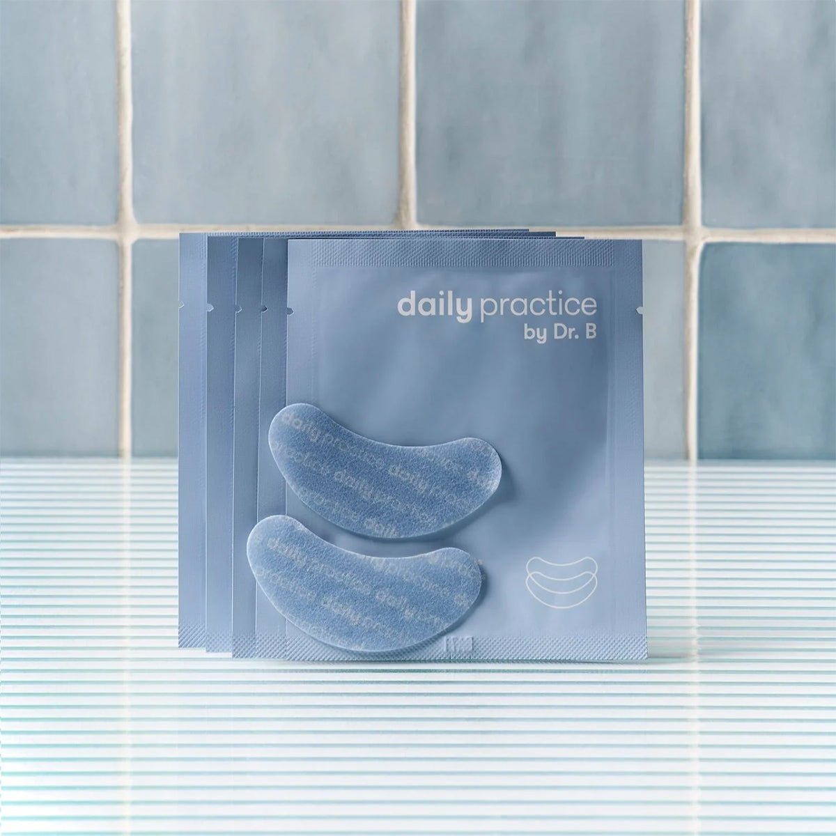 Five blue packets of Daily Practice Brighten & Hydrate Eye Patches – Instantly Refresh & Rejuvenate Your Eyes (5-Pairs) are fanned out on a striped surface, featuring two crescent-shaped patches for reducing puffiness and dark circles. A tiled wall serves as the backdrop.