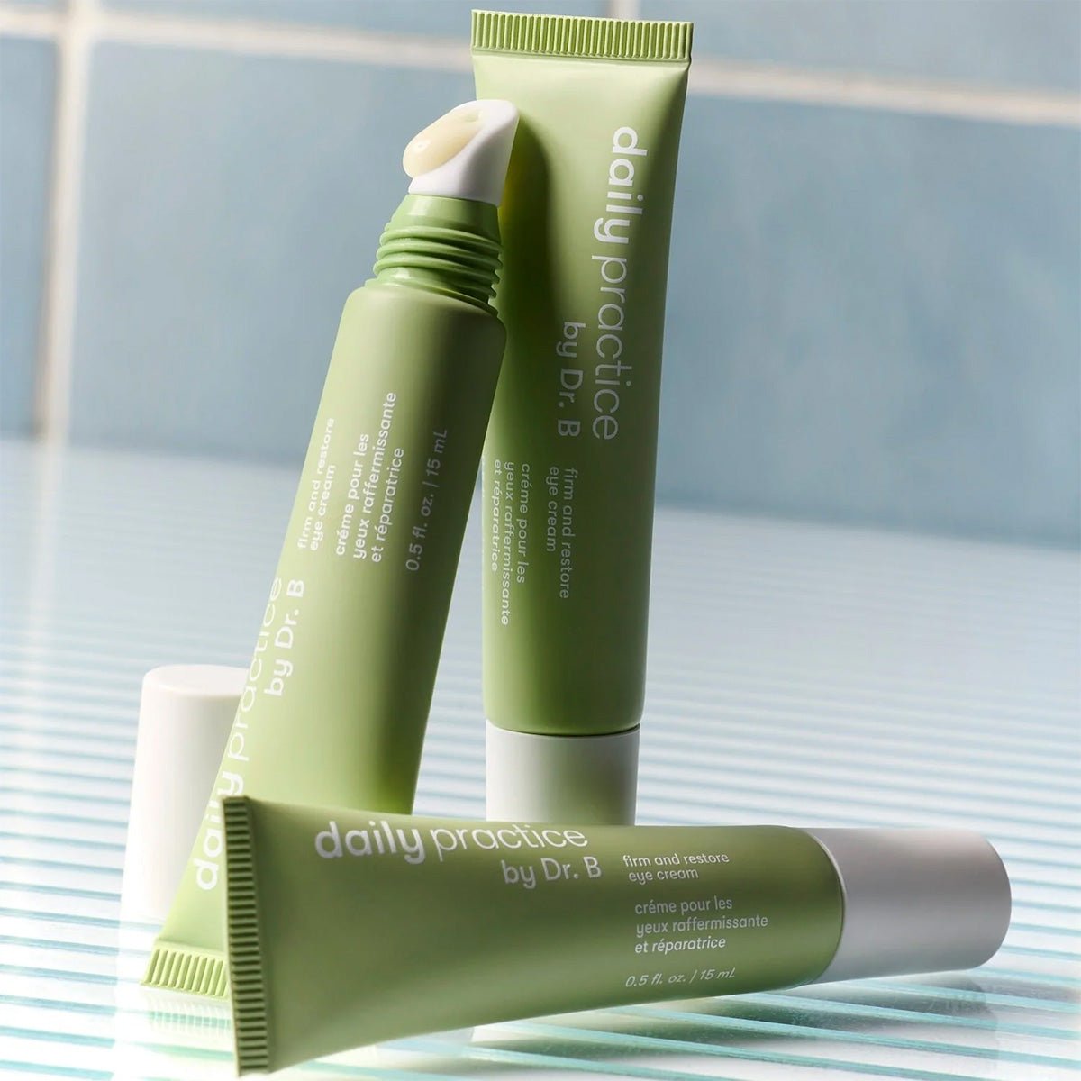 Three green tubes of Daily Practice Firm & Restore Eye Cream – Smooth, Hydrate & Lift (15mL) are shown on a striped surface with a tiled background. One open tube reveals a metal applicator tip for enhancing skin texture and diminishing fine lines.