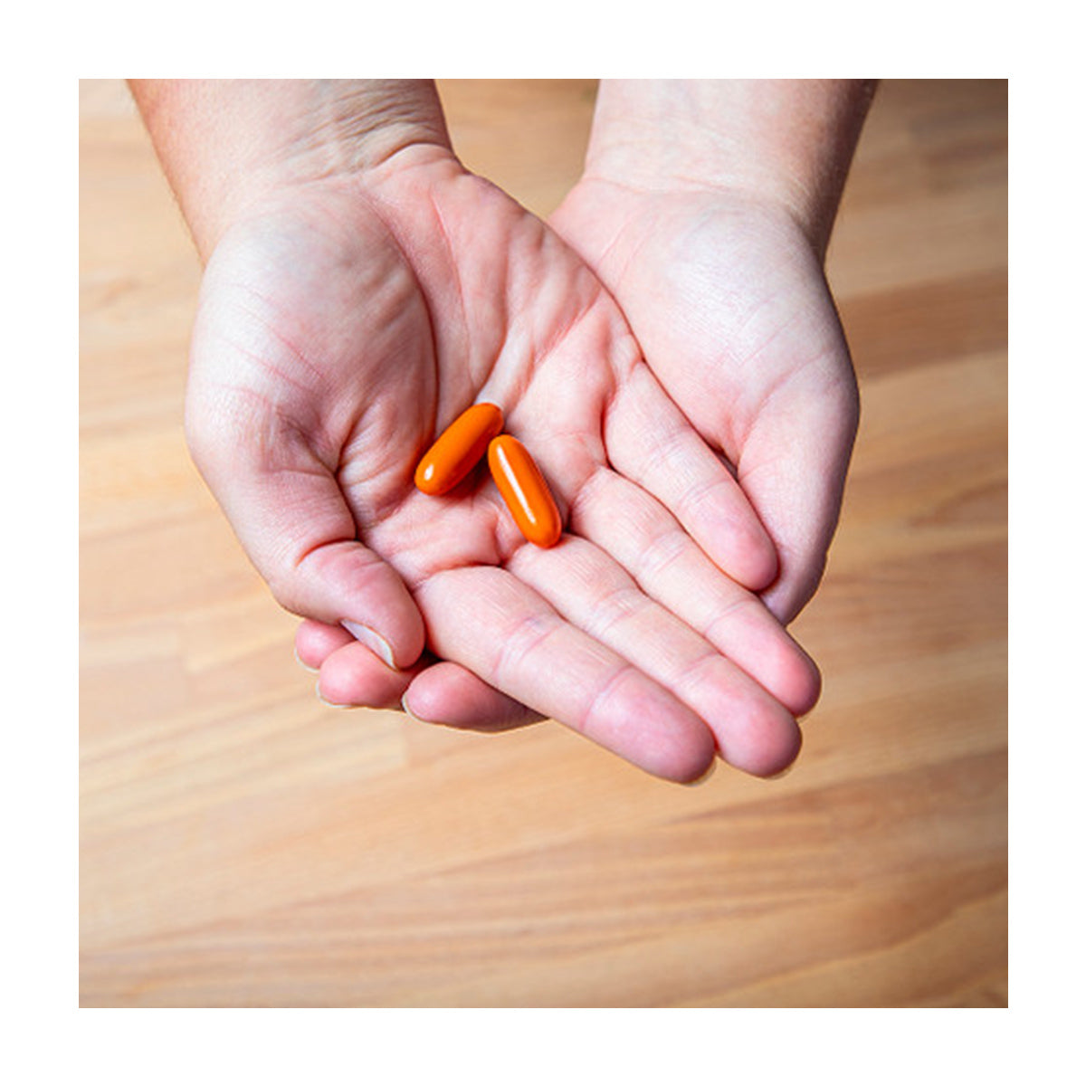 Two hands cup two oval, orange capsules over a wooden floor, representing EyePromise® EZ Tears - Natural Dry Eye Relief with Omega-3s, Turmeric, and Green Tea Extract.