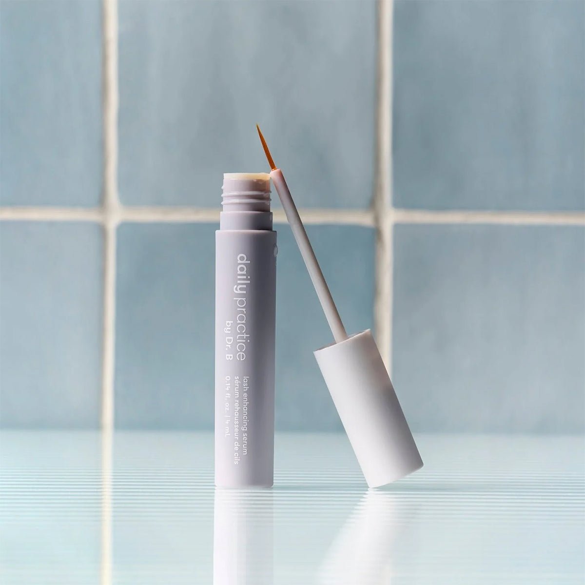 A white tube of Daily Practice Lash Enhancing Serum (4ml) with a thin brush applicator stands open on a reflective surface, set against light blue tiled walls, suggesting fuller, longer, healthier lashes naturally.