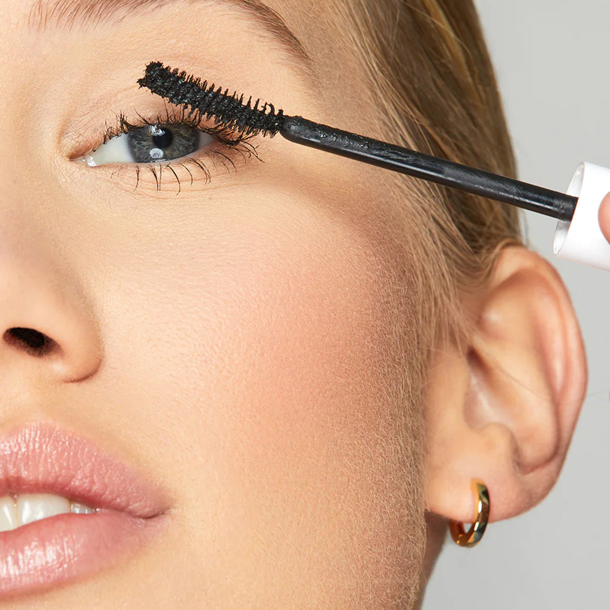 Close-up of a person with light skin applying Twenty Twenty Clean Sweep Mascara in Pitch Black to their upper lashes. The gloss-lipped individual wears small hoop earrings. Ideal for sensitive eyes, the background is plain and neutral.