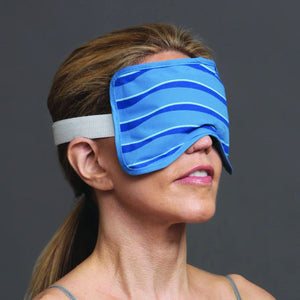 A person with light brown hair wears a Bruder Eye Compress Cold Therapy mask, featuring a blue striped design and a white elastic strap. Set against a plain gray background, their serene expression is highlighted as they face slightly right, perhaps seeking gentle inflammation relief.