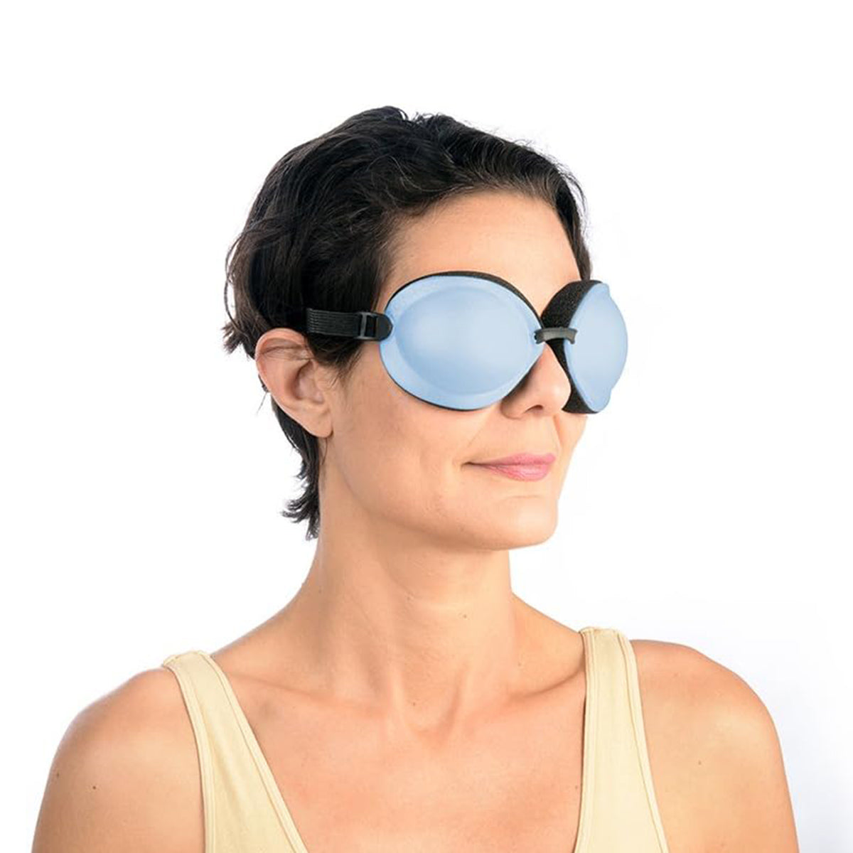 A person with short dark hair and a light beige tank top wears large, opaque blue PRN Tranquileyes XL goggles with a black band against a white background, smiling slightly. They enjoy the soothing dry eye relief from the EyeEco Tranquileyes XL’s moist-heat therapy for severe dry eye and MGD.