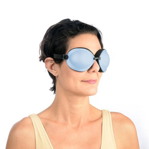 A person with short dark hair and a light beige tank top wears large, opaque blue PRN Tranquileyes XL goggles with a black band against a white background, smiling slightly. They enjoy the soothing dry eye relief from the EyeEco Tranquileyes XL’s moist-heat therapy for severe dry eye and MGD.