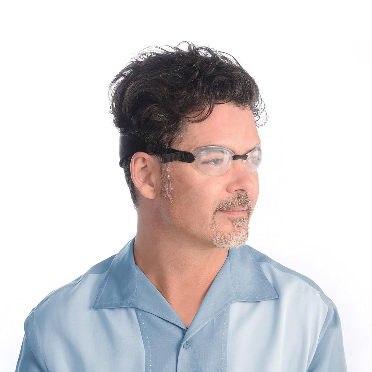 A man with curly hair and a beard wears the EyeEco Quartz Hydrating Sleep Mask from PRN, featuring silicone shields. He is in a light blue collared shirt, gazing to the side against a plain white background.