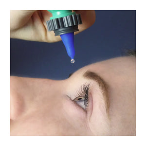 Close-up of a person lying down with their eye open, as a drop from the Nanodropper Eye Drop Extender Adaptor hovers above. The green body and blue nozzle aim to reduce waste against the stark dark background by reducing drops by 60% for easier application.