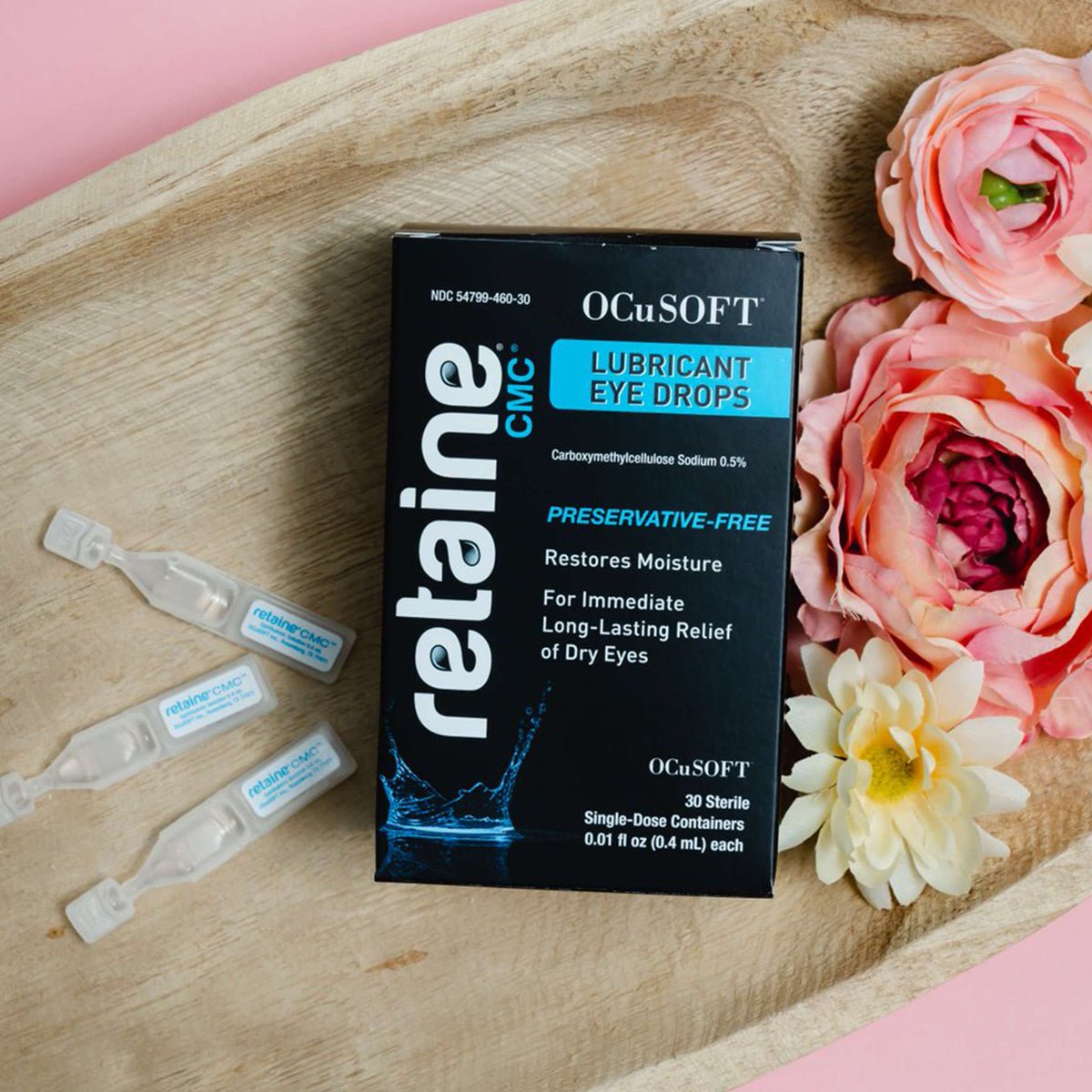 A box of OCuSOFT Retaine CMC Eye Drops, promising preservative-free dry eye relief with 30 single-use vials, rests on a wooden tray adorned with pink and white flowers against a pink background.